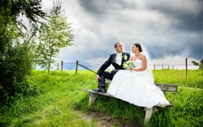 Brautpaar sitzt auf einer Bank, umgeben von gruenen Wiesen in Uffing am Staffelsee, Hochzeitsfotografie im Freien.