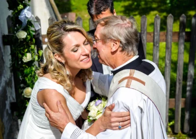 Braut umarmt Priester bei Hochzeitsfeier in Gmund, Hochzeitsfoto mit Blumenstrauss im Hintergrund.