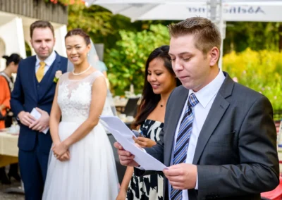 Hochzeitsgast haelt Rede bei Hochzeit nahe Schliersee, Brautpaar im Hintergrund, festlicher Anlass im Freien.