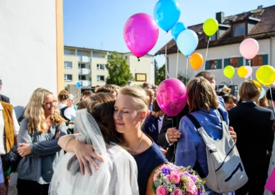 Braut mit Schleier und Blumstrauss umarmt Gast bei kirchlicher Trauung in Grosshadern, bunte Luftballons steigen im Hintergrund.