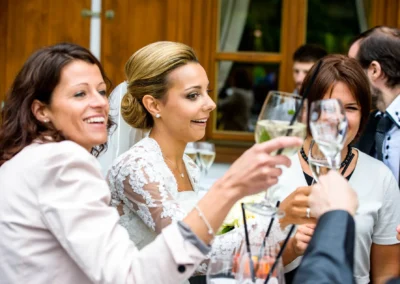 Hochzeitsfoto nahe Tegernsee: Braut im Spitzenkleid mit Gaesten beim Anstossen auf der Hochzeit.