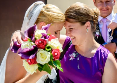 Braut mit Blumenstrauss umarmt lachende Frau bei kirchlicher Trauung in Taufkirchen bei München. Hochzeitsfoto.