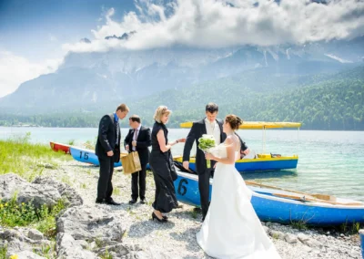 Brautpaar und Gaeste bei der Hochzeit am Eibsee, Berge im Hintergrund, Hochzeitsfotografie unter freiem Himmel.