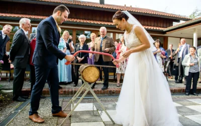 Brautpaar beim Baumsaegen auf Hochzeit nahe Schliersee, Gaeste schauen zu. Hochzeitsfotograf festhaelt Moment.