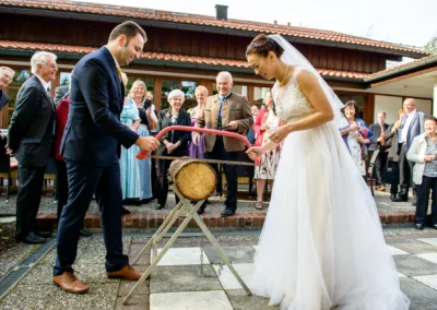 Brautpaar beim Baumsaegen auf Hochzeit nahe Schliersee, Gaeste schauen zu. Hochzeitsfotograf festhaelt Moment.