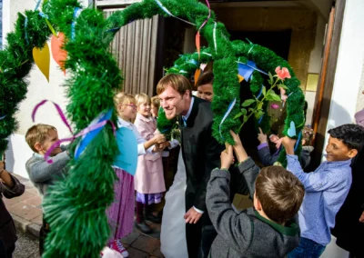 Brautpaar laechelt unter gruenem Kranz mit Kindern, kirchliche Trauung bei Hochzeit nahe Holzkirchen. Hochzeitsfotografie.
