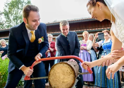 Brautpaar saegt Baumstamm bei Hochzeit nahe Schliersee, Gaeste schauen zu, festgehalten von Hochzeitsfotograf.