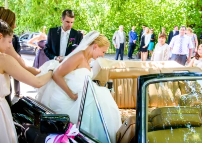 Braut steigt in Auto ein, begleitet von Hochzeitsgesellschaft bei kirchlicher Trauung nahe München. Hochzeitsreportage.