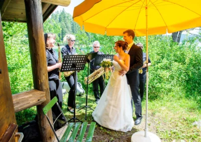 Brautpaar unter gelbem Sonnenschirm mit Musikern im Freien bei Hochzeit nahe Eibsee. Hochzeitsfotografie im Gruenen.