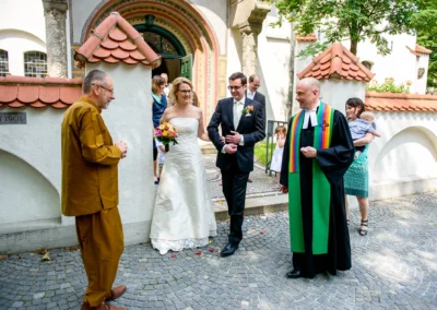 Brautpaar mit Blumenstrauss verlaesst Kirche nach Hochzeit in Schwabing, begleitet von Geistlichem und Gaesten.
