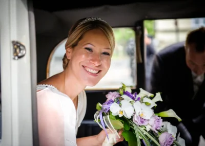 Lächelnde Braut mit Blumenstrauss im Auto, Hochzeitsfotografie in Schwabing, festgehaltene Emotionen der Hochzeit.