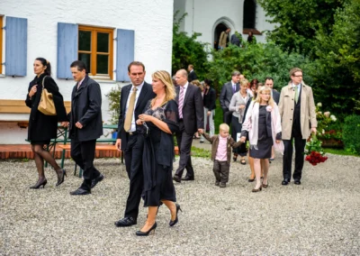 Gäste bei einer Hochzeit in Oberbayern verlassen eine Kirche, Paar mit Rosenstrauss, fotografiert von einem Hochzeitsfotografen.