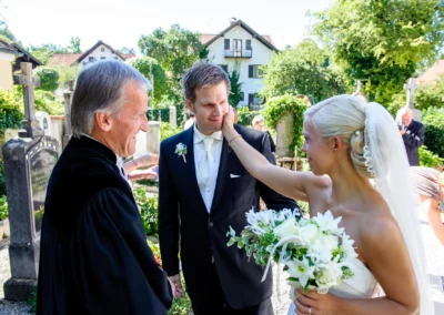 Brautpaar und Geistlicher lachen bei Hochzeit in Bernried am Starnberger See im Freien. Brautstrauss in Nahaufnahme.