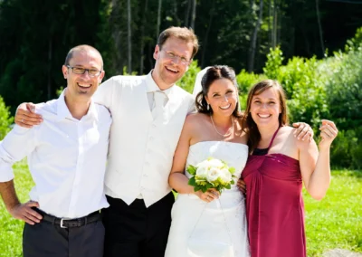 Brautpaar lacht mit Freunden, Sonnenlicht, grüne Bäume im Hintergrund. Hochzeitsbericht auf Sprengenöder Alm unweit von Muenchen.