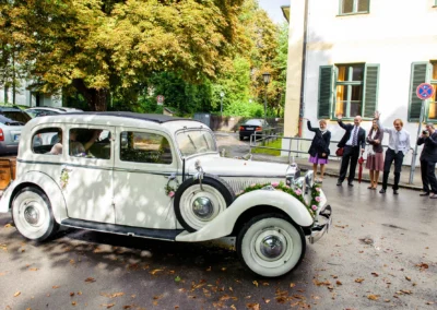 Hochzeitsauto vor historischen Gebaeuden in Schwabing, Gaeste winken zur Feier der Hochzeitsfotografie in Muenchen.