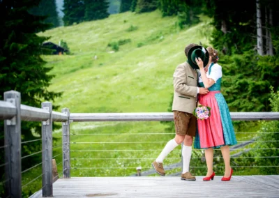 Brautpaar in Tracht auf Holzterrasse, Hochzeitsfoto im Almbad-Sillberghaus, grüne Wiese im Hintergrund.