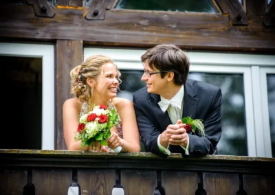 Brautpaar lacht auf Holzbalkon bei Hochzeitsfotografie in Walchensee, Brautstrauss in roten und weissen Blumen.