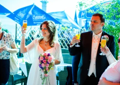 Braut und Bräutigam bei Hochzeitsfeier in Hugos Beach Club mit Blumenstrauß und Gästen im Hintergrund.
