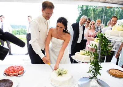 Brautpaar schneidet Hochzeitstorte mit Gaesten im Hintergrund, bei Hochzeit suedlich von Muenchen, von Hochzeitsfotograf.