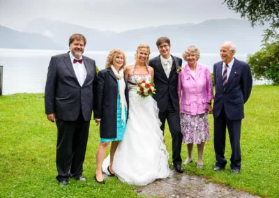 Hochzeitsfoto am Walchensee: Brautpaar und Familie posieren lachend auf einer Wiese mit Bergblick im Hintergrund.
