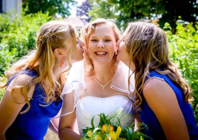 Braut lacht mit Freundinnen in blauen Kleidern im Freien, nahe der evangelischen Friedenskirche bei Starnberg.