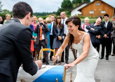 Braut und Bräutigam zersägen ein Holzbrett bei ihrer Hochzeit in Sauerlach-Arget, umgeben von applaudierenden Gästen.