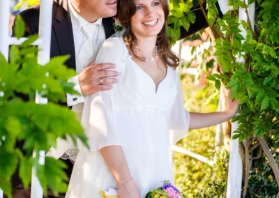 Brautpaar unter Gruen am Hugos Beach Club, laechelnd, Braut mit Blumenstrauss. Hochzeitsfotograf zeigt intime Momente.