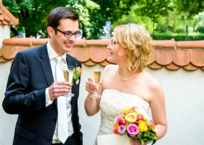 Brautpaar beim Anstoßen mit Sekt in München Schwabing, Hochzeitsfoto mit Blumenstrauss und fröhlicher Stimmung.
