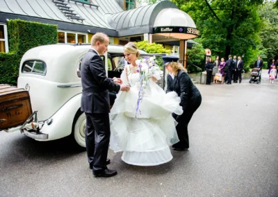 Braut im weissen Kleid steigt aus Oldtimer bei Hochzeit im Seehaus im Englischen Garten in Muenchen. Hochzeitsfotografie.