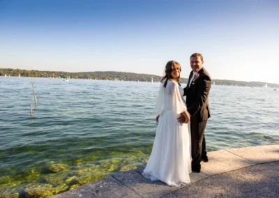 Brautpaar steht am Ufer des Starnberger Sees, umringt von blauem Himmel und Segelbooten. Hochzeitsfoto im Freien.