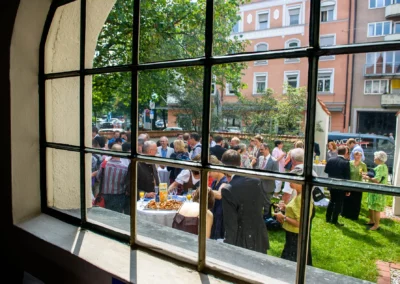 Hochzeitsreportage in Schwabing: Gäste draußen im Garten bei Sektempfang, durch Fenster fotografiert.