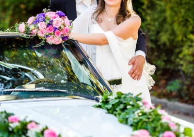 Brautpaar steht lächelnd vor geschmücktem Auto bei Hochzeit am Starnberger See, Hochzeitsfotograf hielt den Moment fest.