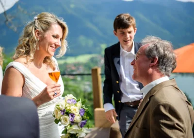 Braut bei Hochzeit im Leeberghof Tegernsee, laechelnd mit Blumenstrauss und Glas, Hochzeitsfotografie im Freien.
