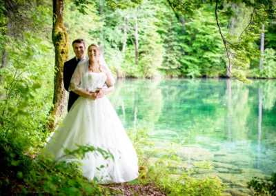 Brautpaar steht am See bei Grainau, umgeben von gruenen Baeumen. Hochzeitsfoto von einer romantischen Hochzeit.