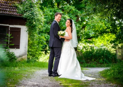Brautpaar laechelnd im Park bei der Hochzeitsfotografie in Oberbayern, Portraitaufnahme neben einem alten Haus.