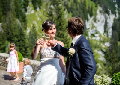 Brautpaar stößt mit Sekt an, umgeben von Natur. Kinder spielen im Hintergrund. Hochzeit bei Kampenwand.