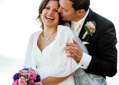 Brautpaar lachend bei Hochzeit am Starnberger See, der Bräutigam küsst die Braut, beide in festlicher Kleidung.