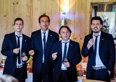 Vier Maenner in Anzuegen bei einer Hochzeit in Arget, trinken Wein und halten Reden vor Holzwand. Hochzeitsreportage.