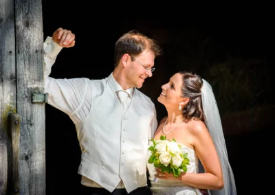 Brautpaar bei Hochzeitsfotografie in Wolfratshausen, er helles Gilet, sie Halsschmuck, lachend mit Blumenstrauss.