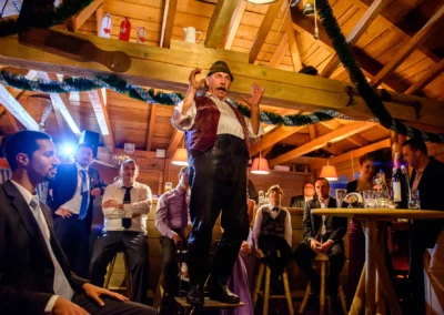 Hochzeitsreportage in Paulaner Seegarten: Ein Mann in traditioneller Kleidung spricht vor sitzenden Hochzeitsgaesten.