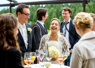 Brautpaar lacht mit Gaesten bei Hochzeit in Waakirchen, Hochzeitsreportage mit Blumenstrauss und Getraenken im Freien.