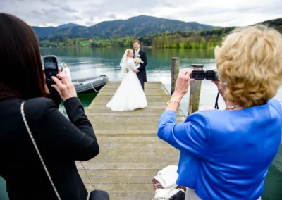 Brautpaar auf Steg am Tegernsee, fotografiert von Gästen. Hochzeitsaufnahme in malerischer Kulisse.