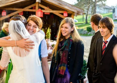 Braut umarmt Gast bei Hochzeit in Bruckmuehl, lachende Freunde beobachten. Hochzeitsreportage im Freien.