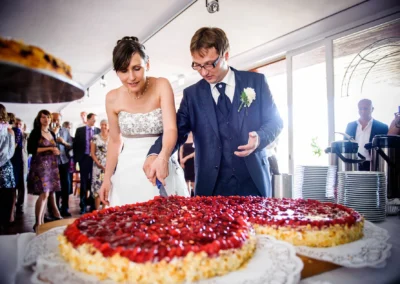 Brautpaar schneidet Hochzeitstorte bei Feier im Chiemgau mit Gästen im Hintergrund. Hochzeitsfotografie.
