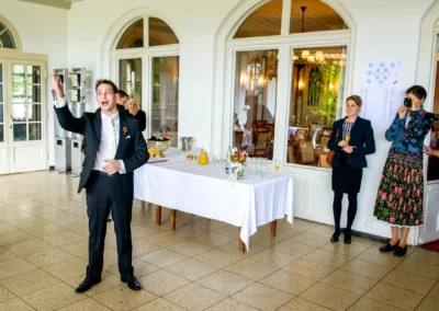 Mann in Anzug haelt Rede bei Hochzeit im Hotel Kaiserin Elisabeth. Drei Personen im Hintergrund neben Tisch.