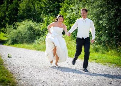 Brautpaar lacht und rennt auf Schotterweg, Hochzeitsreportage bei Wolfratshausen, Hochzeitsszene in der Natur.
