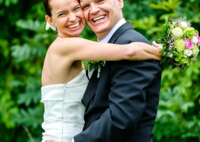Glueckliches Paar bei Hochzeit auf Gut Thurnsberg. Frau im Brautkleid umarmt Bräutigam, lacht und hält Blumenstrauss.