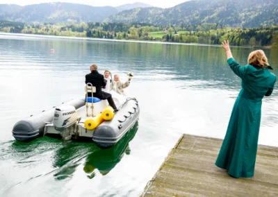 Brautpaar auf Boot bei Hochzeit in Gmund am Tegernsee, Frau am Steg winkt. Romantische Hochzeitsfotografie in Bayern.