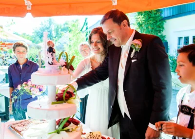 Brautpaar schneidet Hochzeitstorte bei Feier in Hugos Beach Club. Hochzeitsfoto zeigt festlichen Moment im Freien.