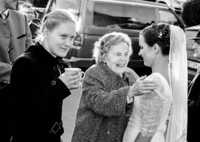 Ältere Frau umarmt Braut bei einer Hochzeitsreportage am Hofgut zur Muehle. Weitere Gaeste im Hintergrund.
